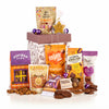 A curated gift basket of assorted snacks and chocolates from brands like Mighty Fine, Elizabeth's, and Popcorn Snaps, arranged around a patterned gift box on a white background.