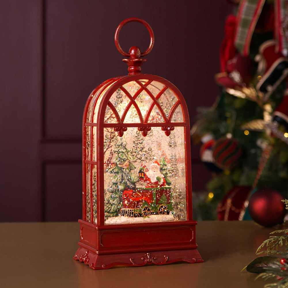 A red arched lantern-style Christmas ornament with a Santa Claus and sleigh scene inside, displayed on a tabletop with a blurred Christmas tree in the background.