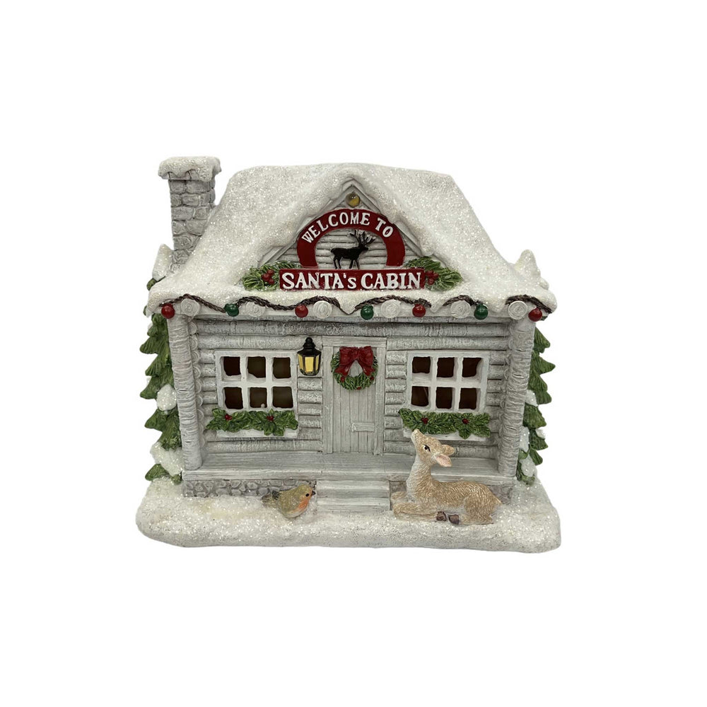 A small Santa's Cabin Christmas ornament with snow-covered gray siding, a lit window, wreath, bird feeder, and a deer on the porch, shown on a plain white background.