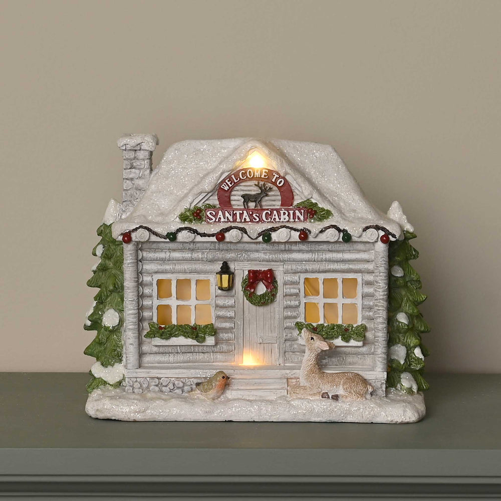 A decorative Santa's Cabin Christmas ornament with a snowy log cabin design, warm glowing windows, a wreath, birds, and a deer, displayed on a shelf against a neutral background.