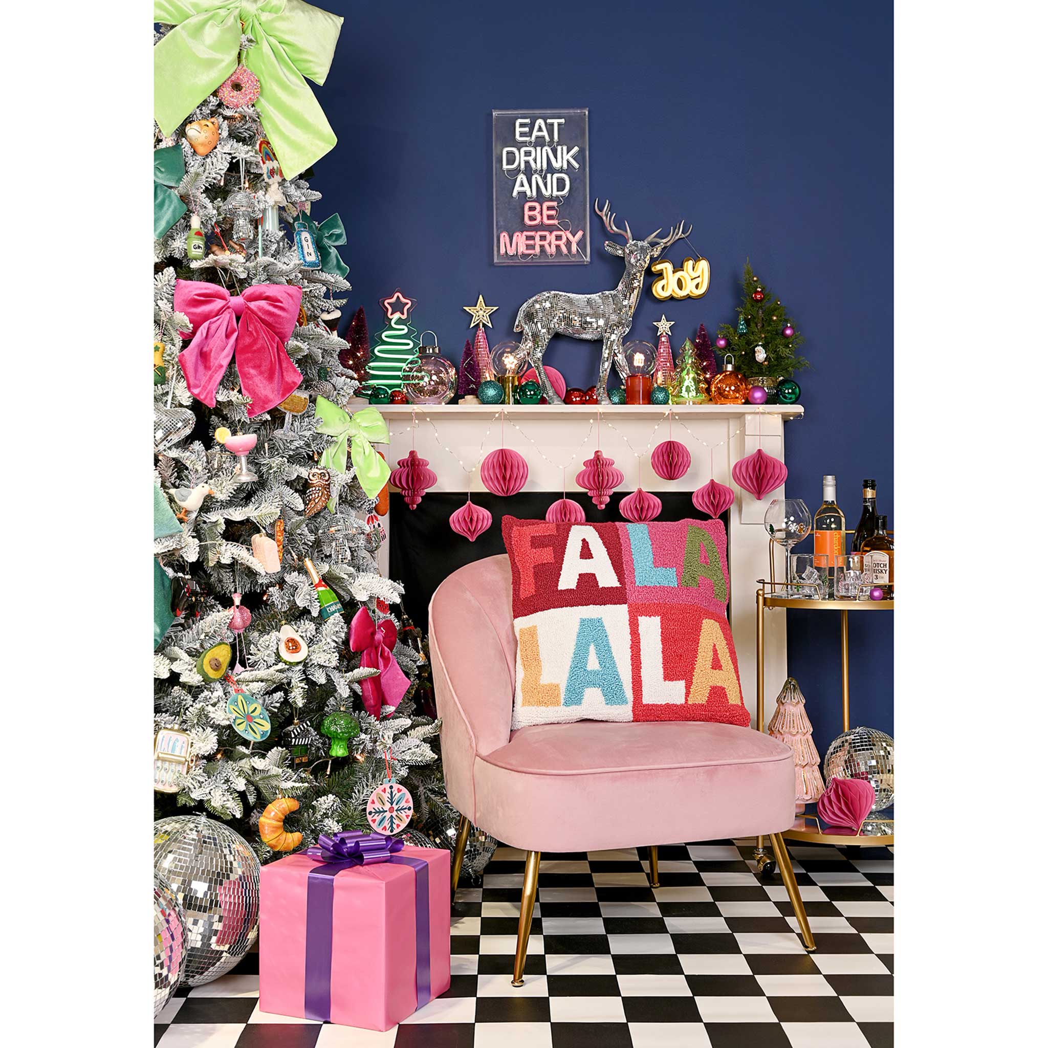 A festive holiday interior scene with a decorated silver Christmas tree, pink armchair with a colorful 'FALA LALA' throw pillow, pink gift box, fireplace mantel with holiday decor, and a dark blue wall.
