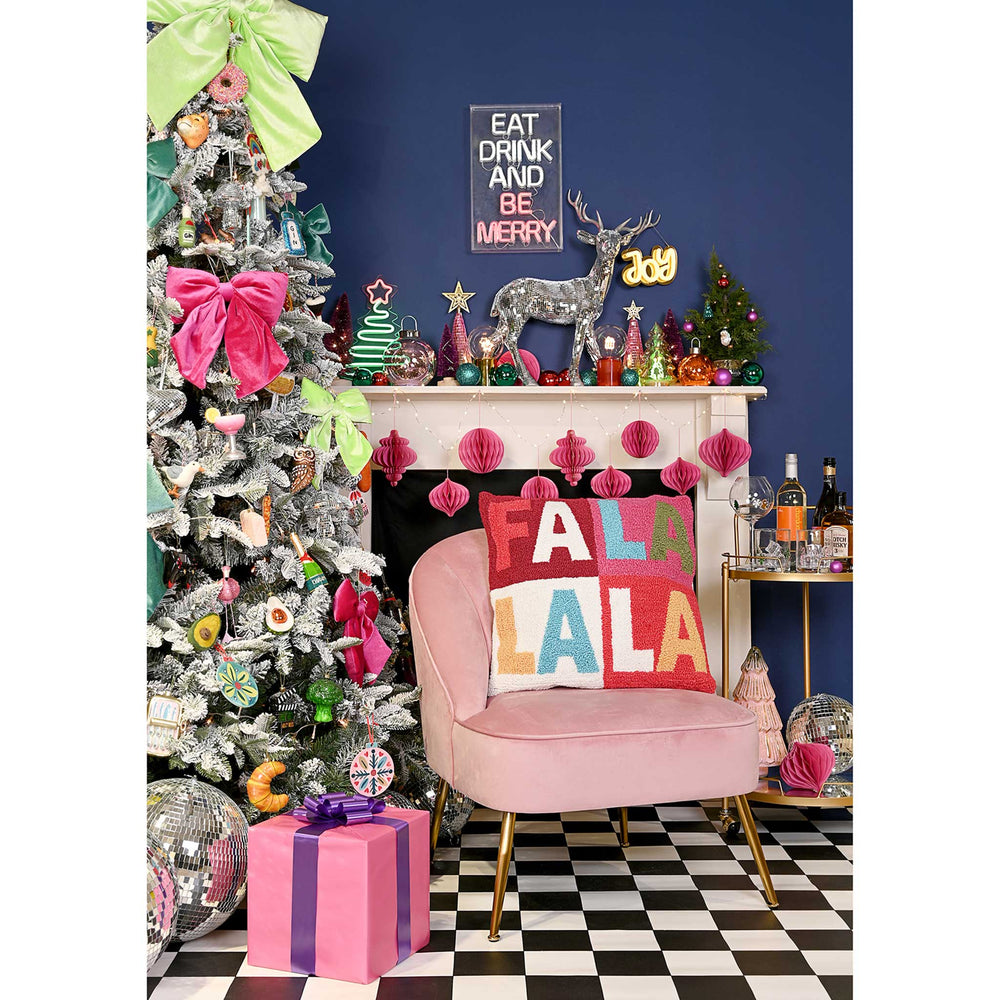 A festive holiday interior scene with a decorated silver Christmas tree, pink armchair with a colorful 'FALA LALA' throw pillow, pink gift box, fireplace mantel with holiday decor, and a dark blue wall.