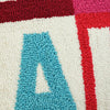 Close-up of a pillow showing a large turquoise letter A on an off-white background, bordered by red and magenta sections.