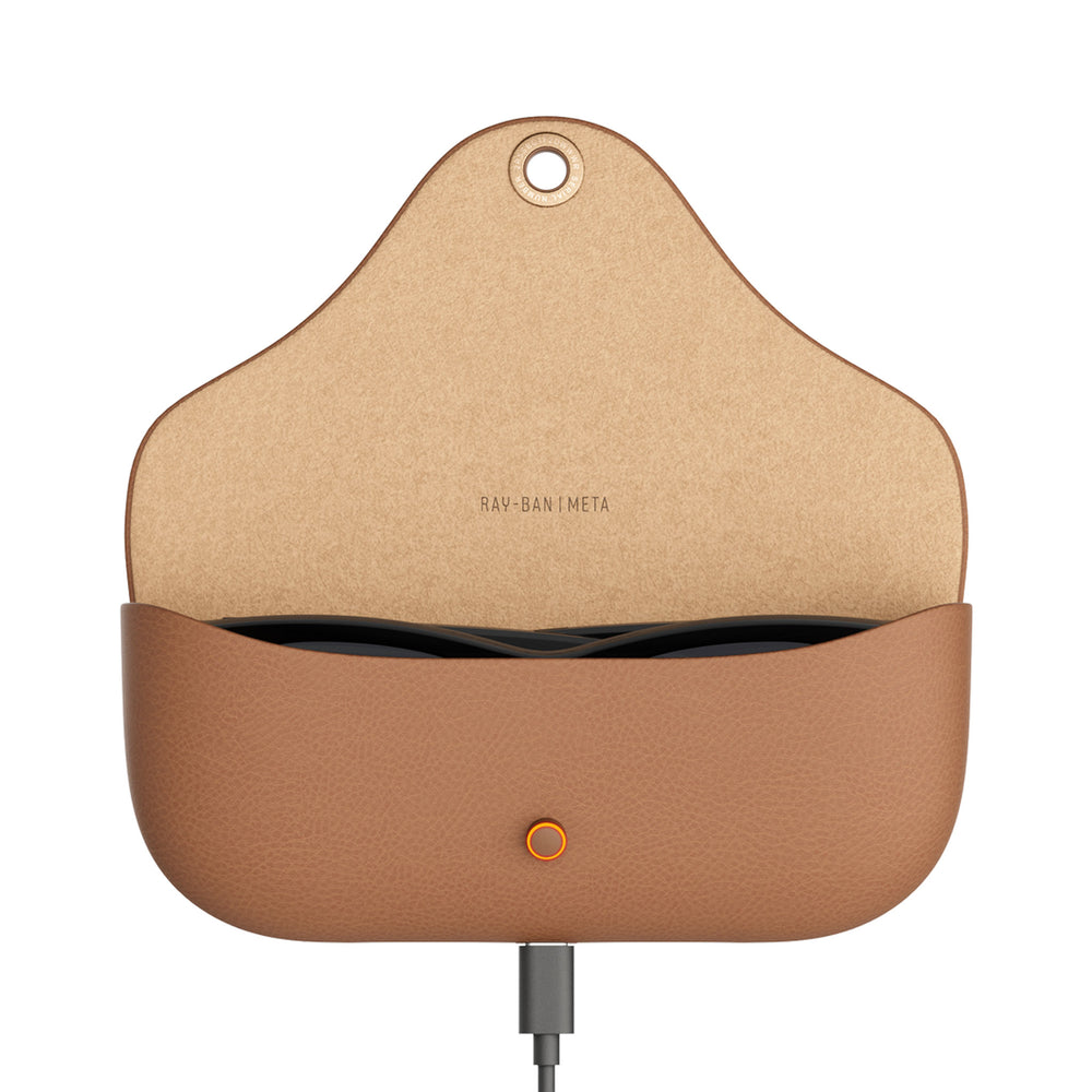 A tan leather charging case for Ray-Ban Meta earbuds, shown open with the earbuds inside and a charging cable plugged into the bottom port. The interior is a natural tan color with the brand name 'RAY-BAN META' printed on it.