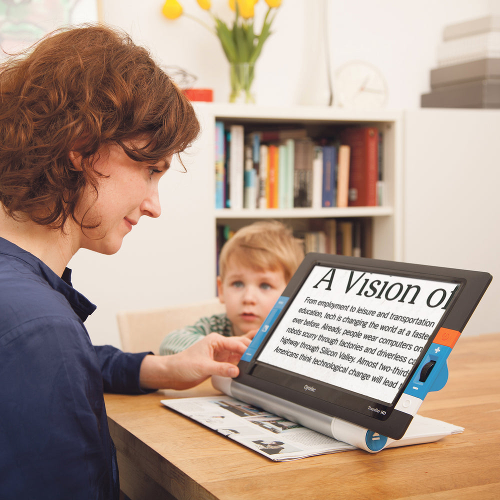 A woman sitting at table reading a newspaper with the traveller HD portable video magnifier