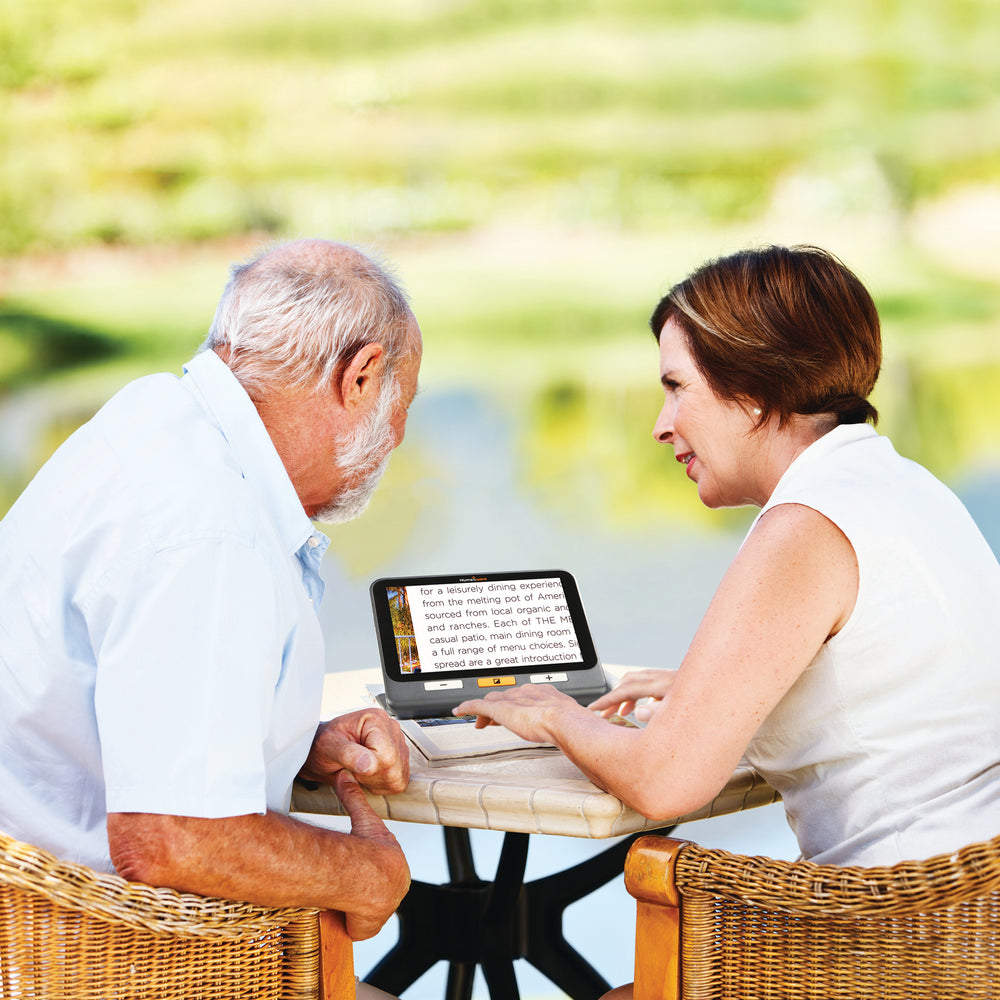 A white man and white woman sit in wicker chairs using the magnifer to read a magazine.