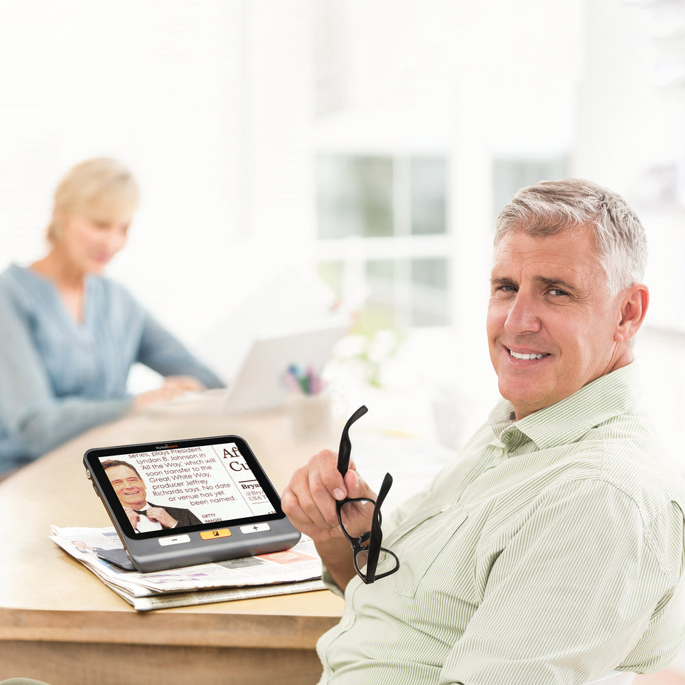 A middle-aged white man looks at the camera while the magnifier sits near him, showing an item in a newspaper. A white woman is out of focus in the background.