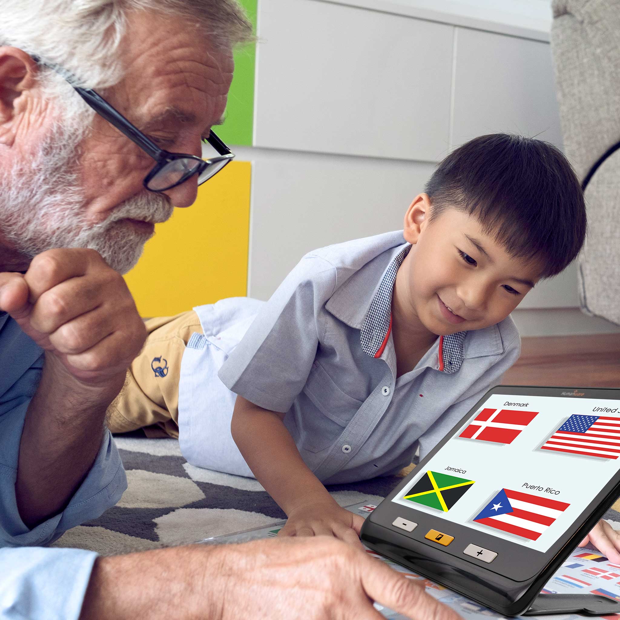 An older man and his grandson lie on the floor and use the Explore 12 to magnify flags of the world