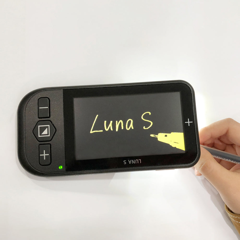 A person using the Luna S video magnifier on its stand to write the words 'Luna S' with a pen. The screen is using yellow on black format