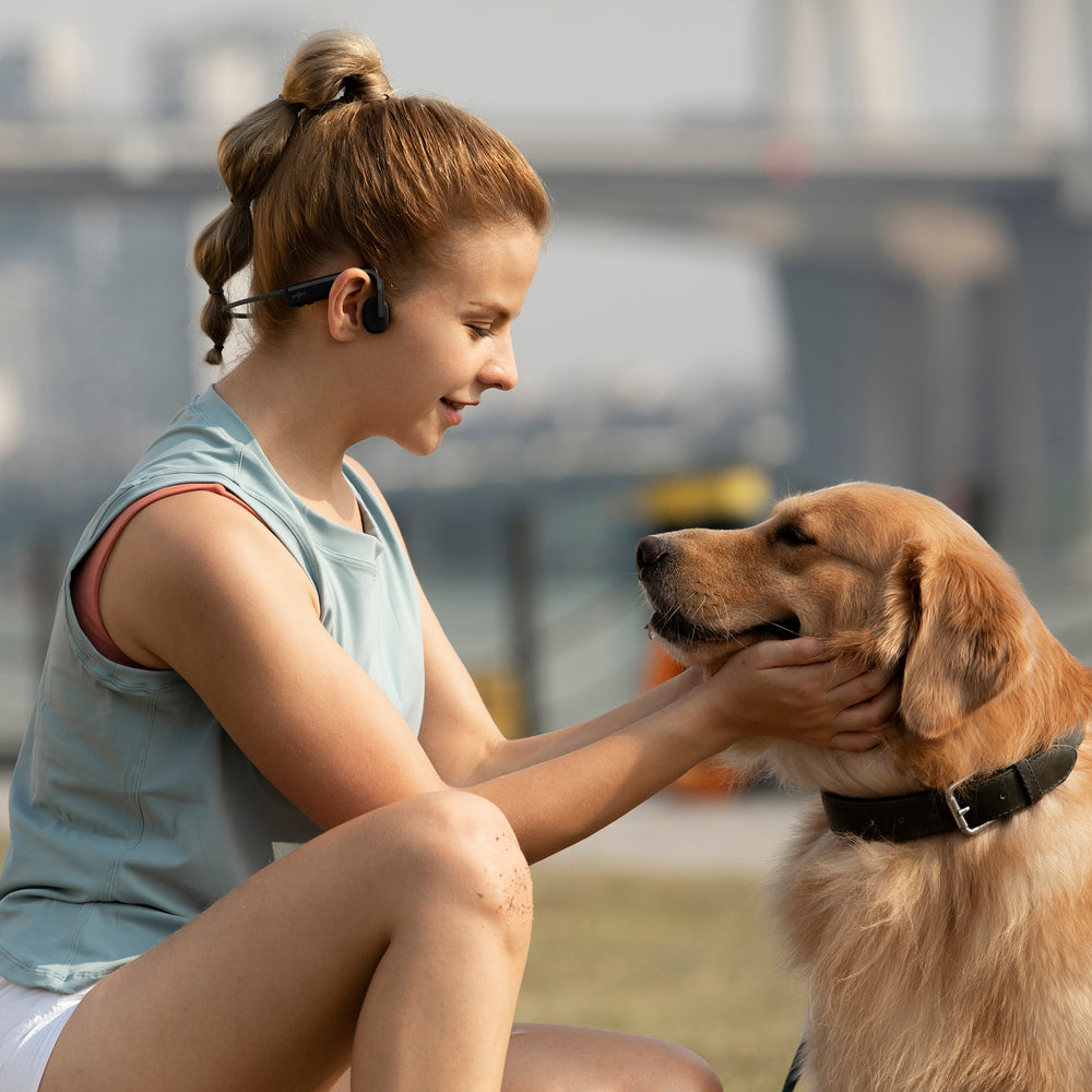 Woman wearing OpenMove Shokz in park petting a dog.