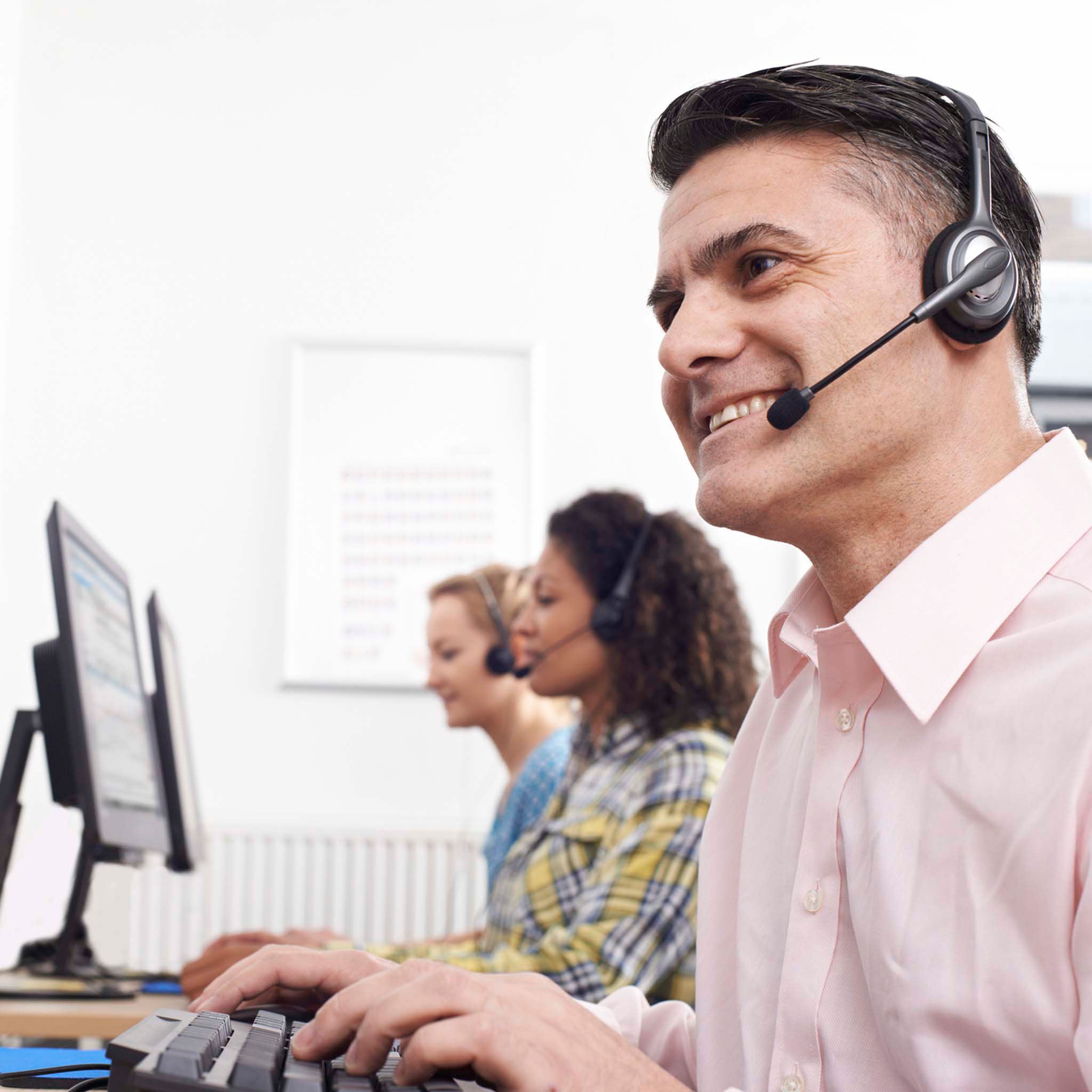 A man in a pink shirt wearing a headset and typing on a computer keyboard in a call center, with two other agents blurred in the background.