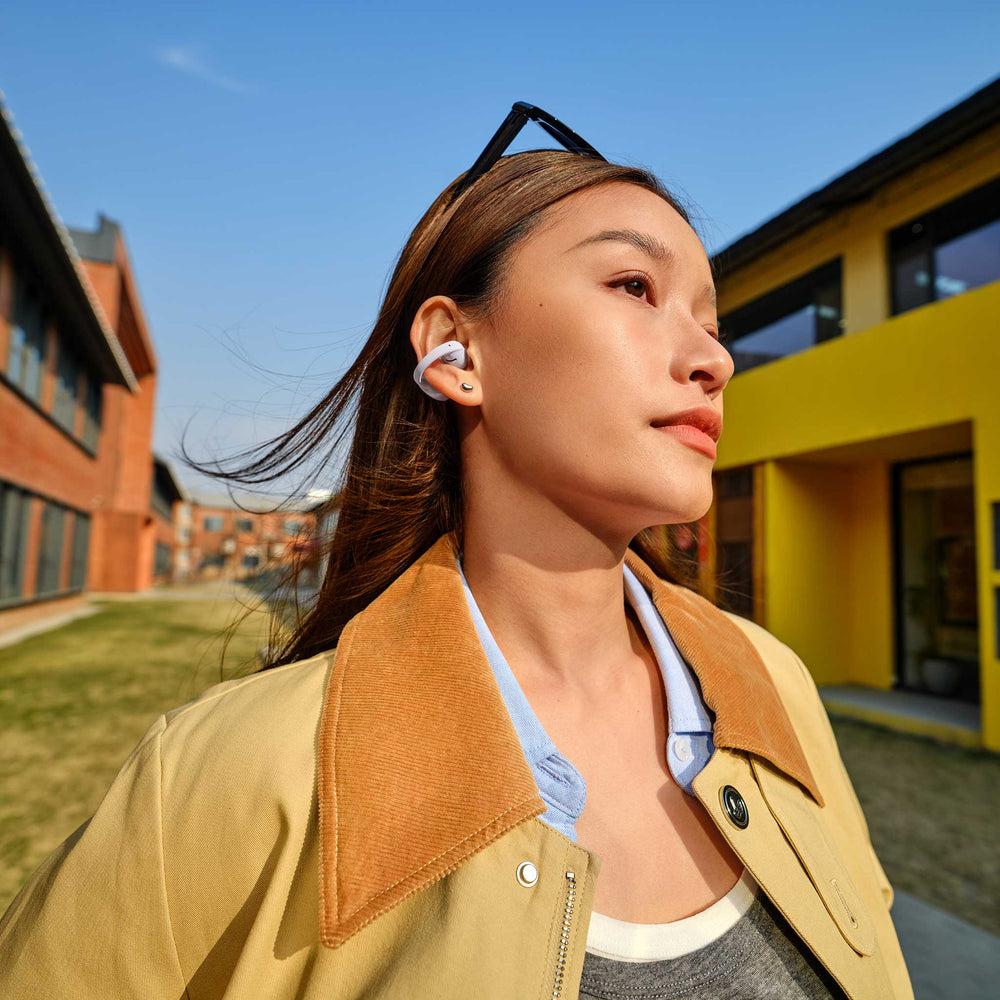A woman outdoors wearing a white wireless earbud, dressed in a beige jacket with a brown suede collar over a light blue shirt. The background features modern buildings under a clear blue sky.