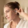 A woman wearing a white collared shirt inserts a small white in-ear earbud into her ear. The background is a softly blurred warm beige indoor setting.