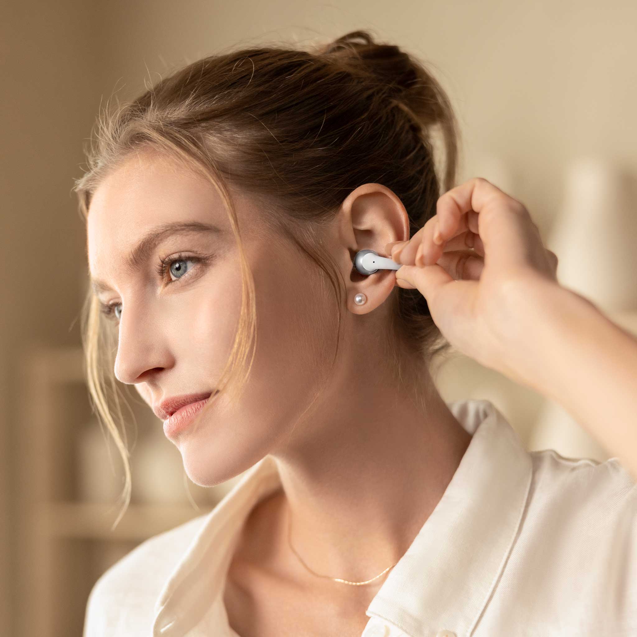 A woman wearing a white collared shirt inserts a small white in-ear earbud into her ear. The background is a softly blurred warm beige indoor setting.