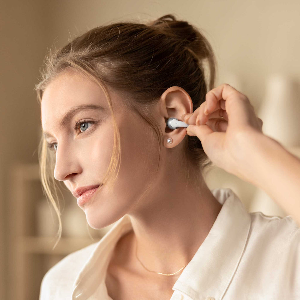 A woman wearing a white collared shirt inserts a small white in-ear earbud into her ear. The background is a softly blurred warm beige indoor setting.