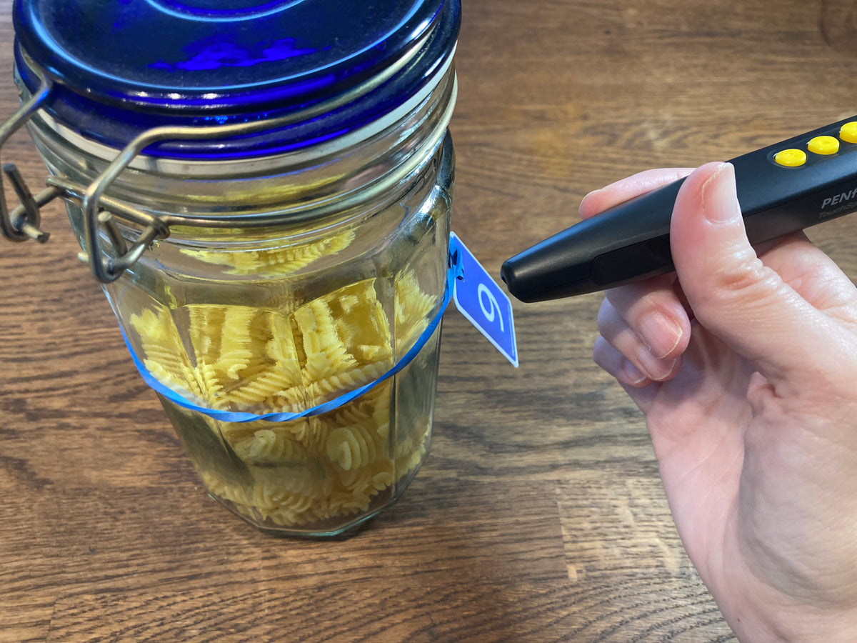 A person using an RNIB Penfriend to read a numbered tag attached to a glass jar containing pasta by an elastic band.