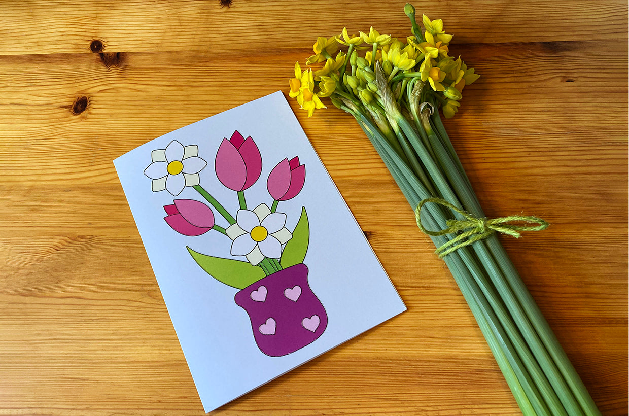 Tactile Mother's Day card featuring a vase containing tulips and narcissi lying on a wooden table next to a bunch of yellow narcissi tied with green twine