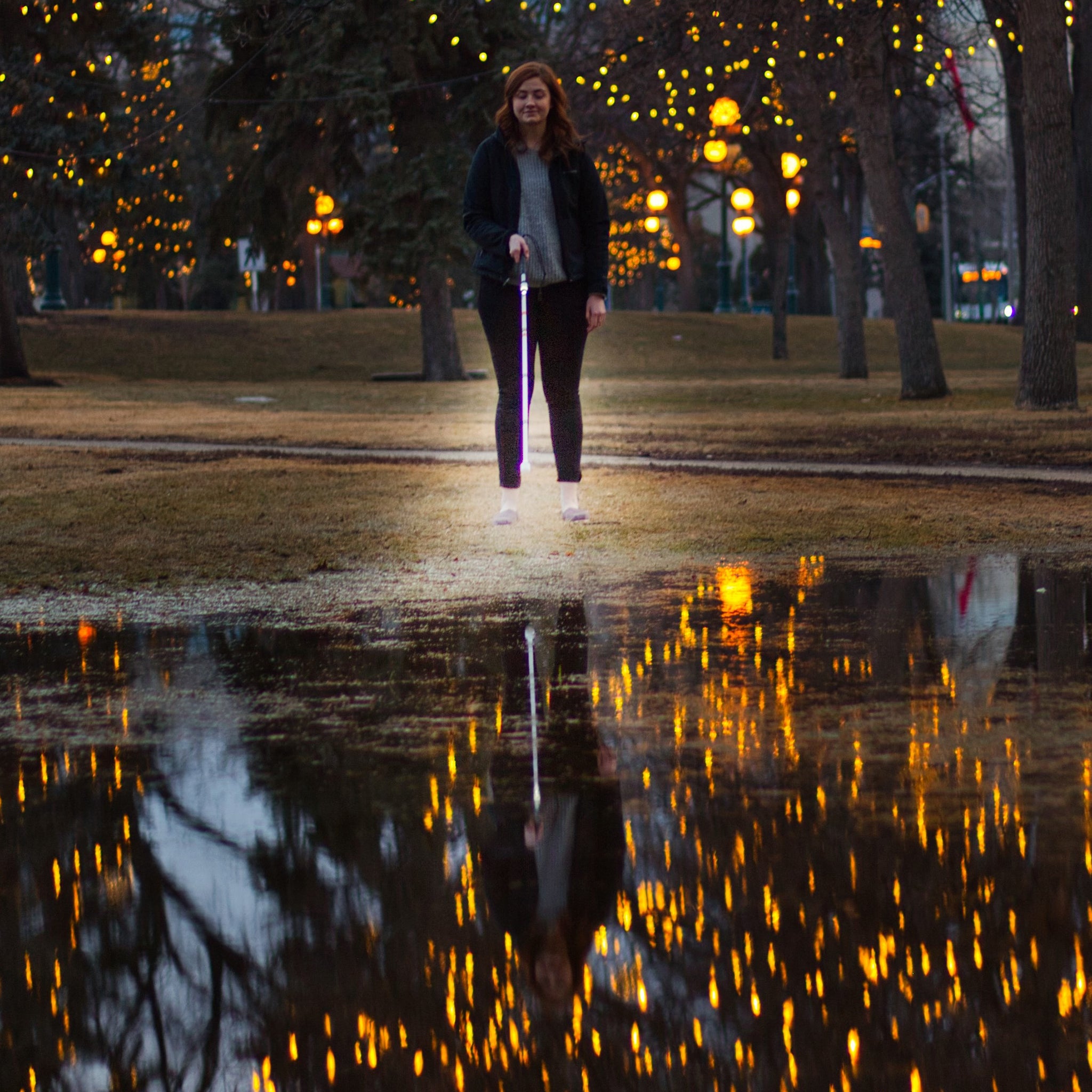 Woman with a cane standing in front of a large puddle with yellow lights strung in trees. The cane light and yellow lights reflect in the puddle.
