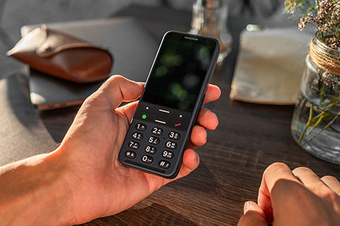 A blue BlindShell Classic 3 smartphone held in a person's hand over a wooden table.