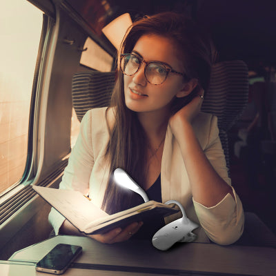 A young woman on a train reading her book with the light clipped to it