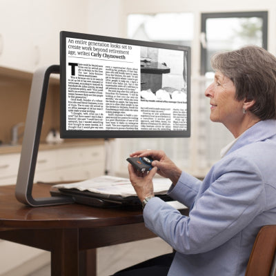 A woman sitting at a table reading a magazine with the ClearView C 24"" HD Desktop Video Magnifier