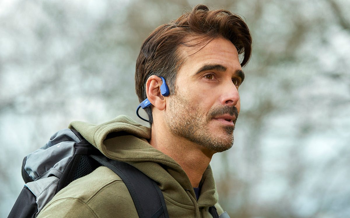 A man with dark hair and short beard wearing an olive green hoodie and backpack listening to audio on blue bone-conducting headphones