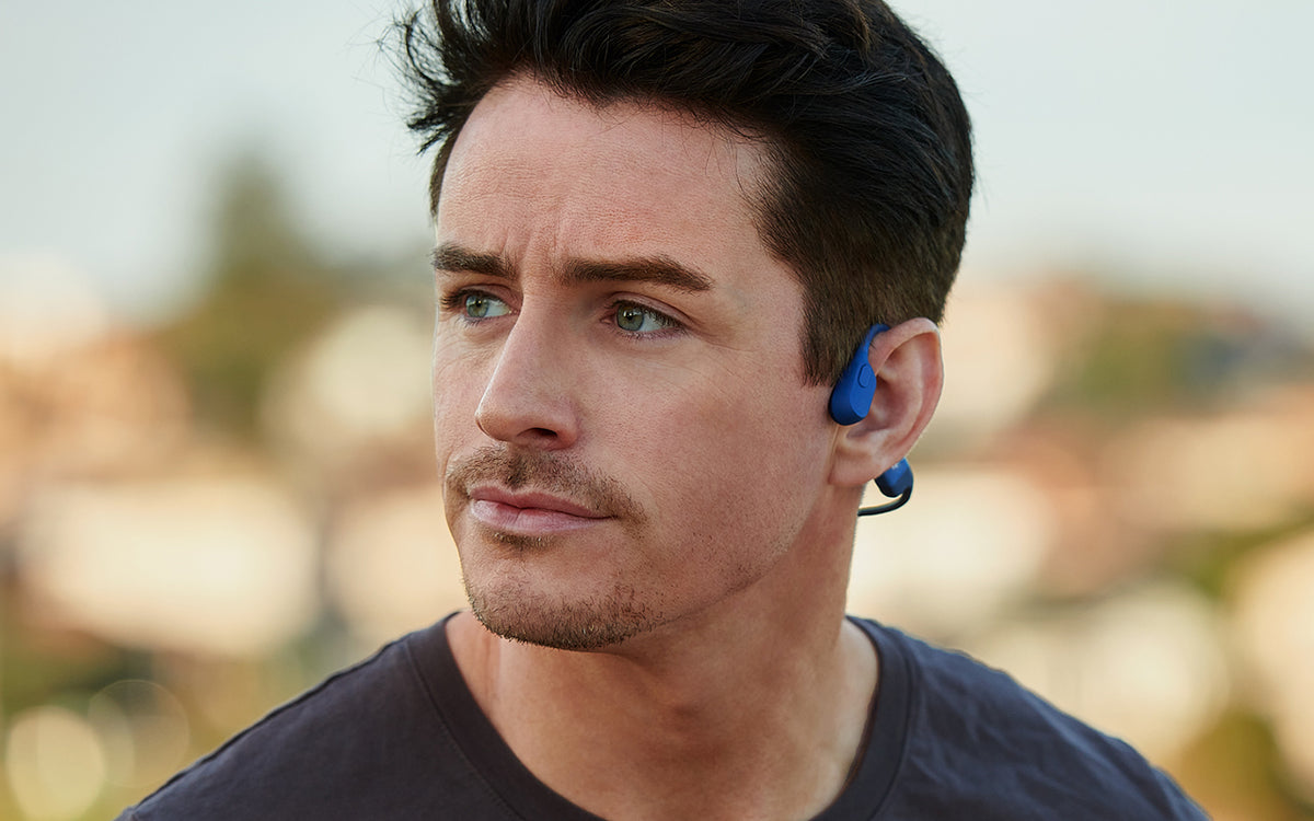 A white man with dark hair and stubbly facial hair looking away from the camera while wearing blue bone-conducting headphones.