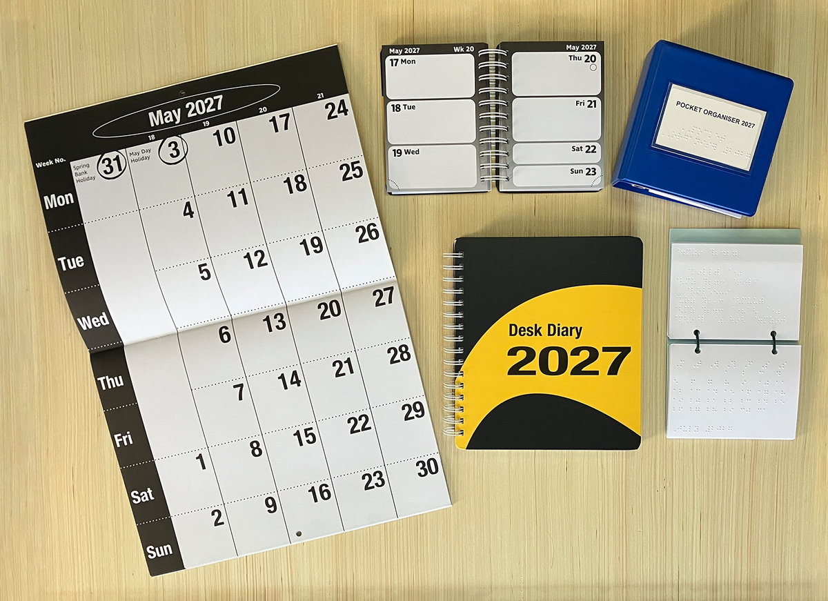 A selection of large print and braille diaries and calendars arranged on a bamboo sheet. The large print calendar, large print pocket diary and braille pocket calendar are open to show the layout inside. The large print desk diary and braille pocket organiser are closed.