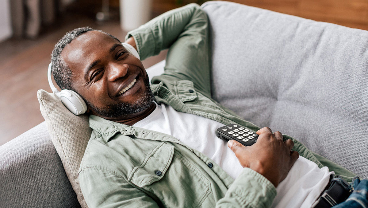 A black man with short salt and pepper hair and beard lies on a sofa smiling and listening to audio on wireless headphones from a portable DAISY player