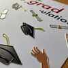 Close-up of a graduation-themed card featuring metallic foil cutouts of graduation caps, diplomas, and a hand.