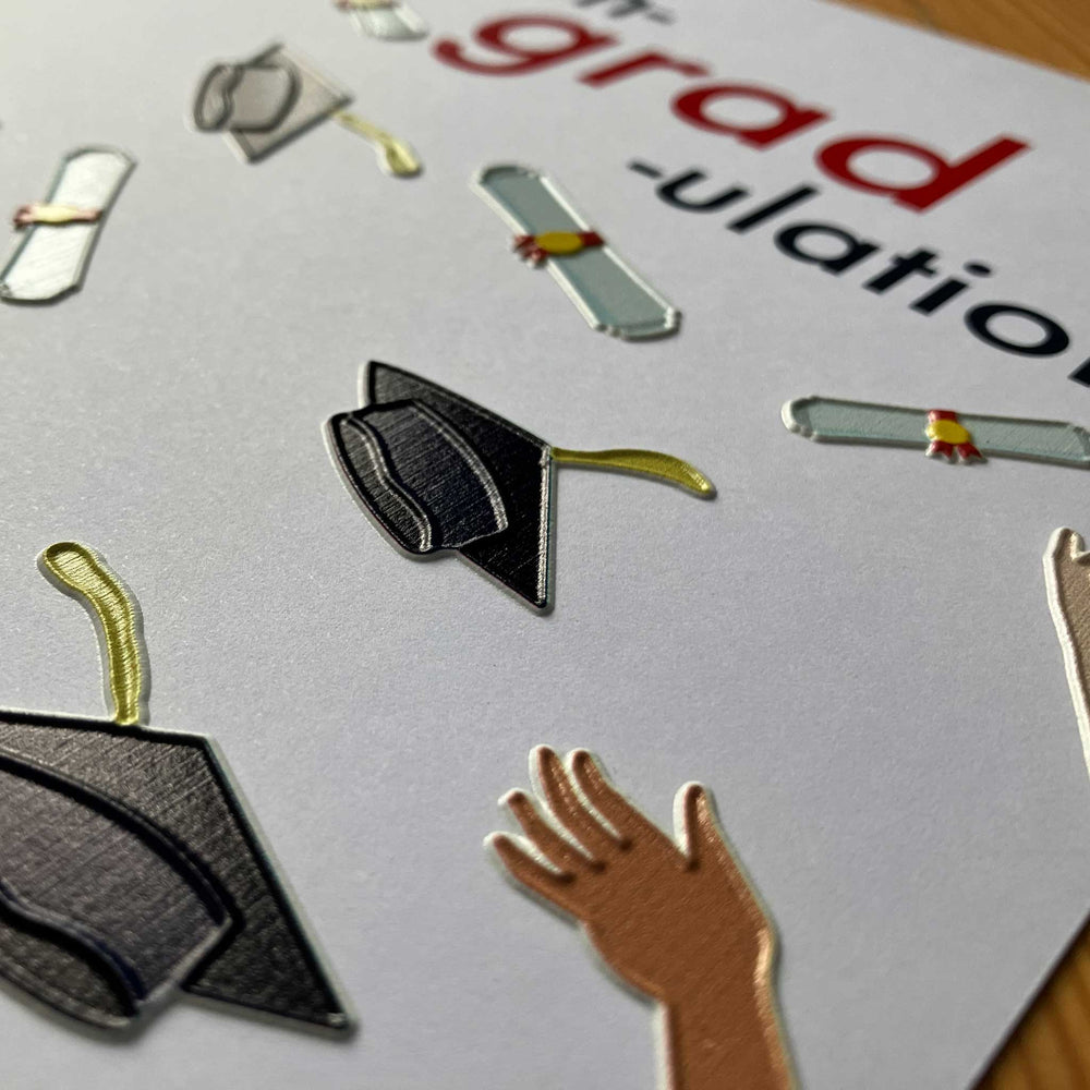 Close-up of a graduation-themed card featuring metallic foil cutouts of graduation caps, diplomas, and a hand.