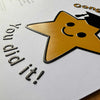 A close-up of card featuring a large gold star with a black outline, a simple smiley face, and part of a black graduation cap. The text 'You did it!' appears in raised dark lettering on the left, with additional gold text partially visible at the top right.