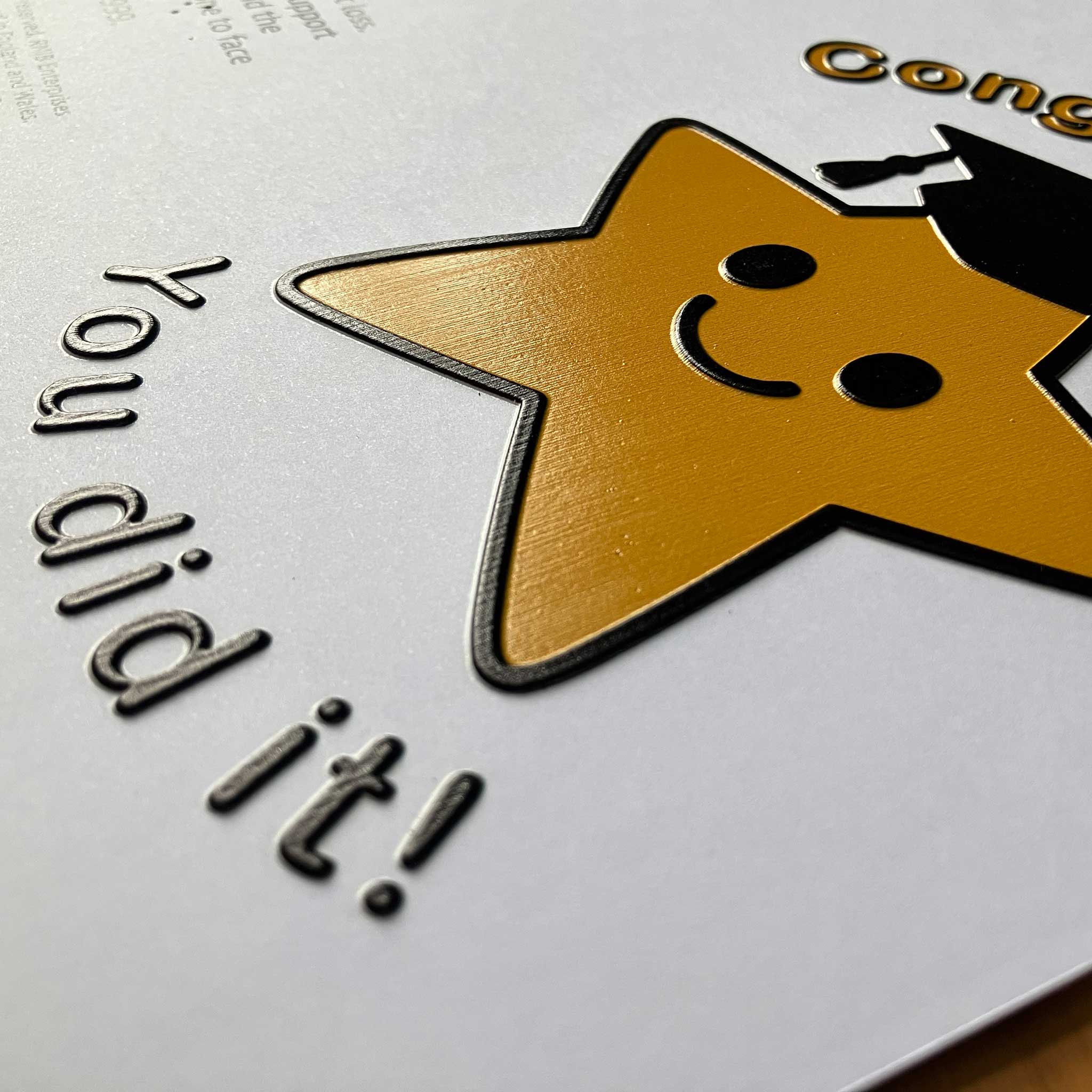 A close-up of card featuring a large gold star with a black outline, a simple smiley face, and part of a black graduation cap. The text 'You did it!' appears in raised dark lettering on the left, with additional gold text partially visible at the top right.