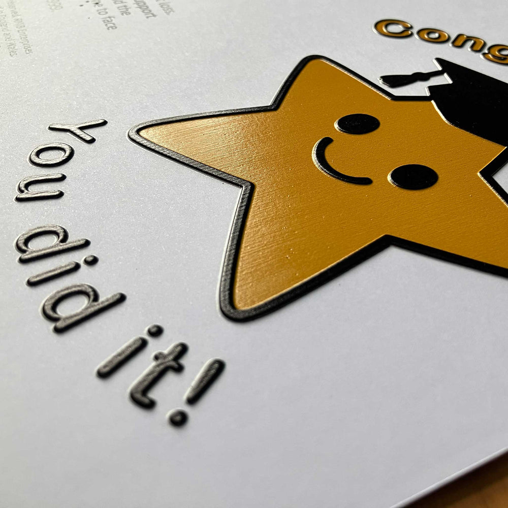 A close-up of card featuring a large gold star with a black outline, a simple smiley face, and part of a black graduation cap. The text 'You did it!' appears in raised dark lettering on the left, with additional gold text partially visible at the top right.