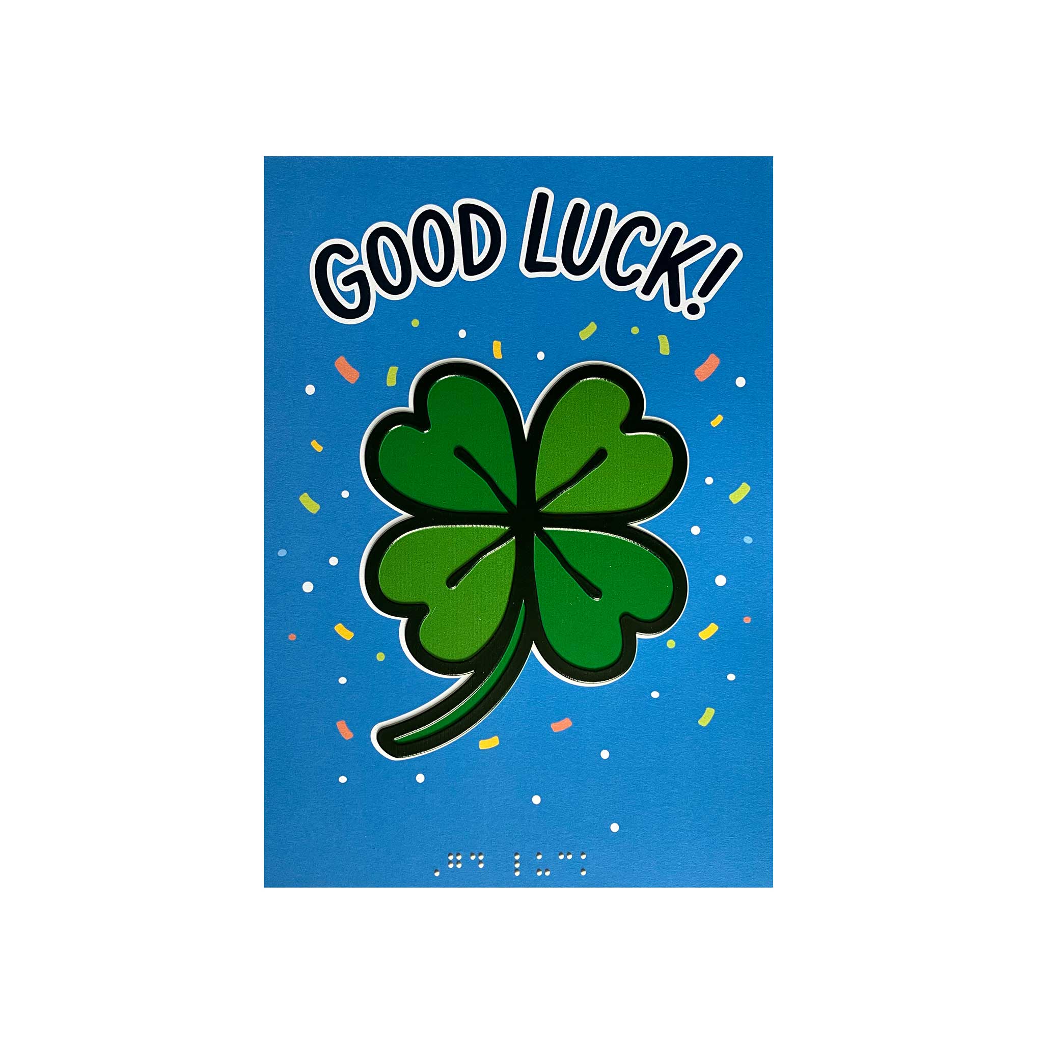 A blue greeting card featuring a large green four-leaf clover with the words 'GOOD LUCK!' in white outlined lettering at the top. Small multicolored confetti dots are scattered around the design, and braille text is visible near the bottom.