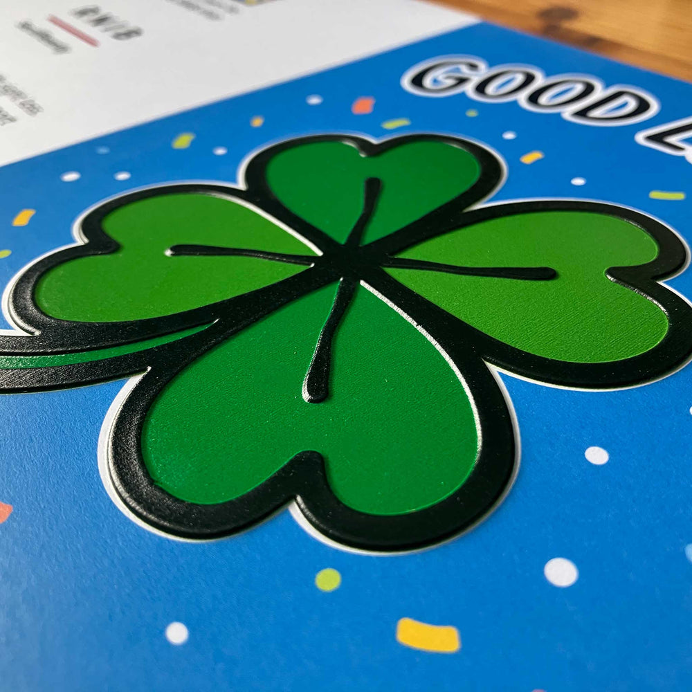 A close-up of a green four-leaf clover sticker or decal with black outlines, applied to a blue printed surface with confetti dots.