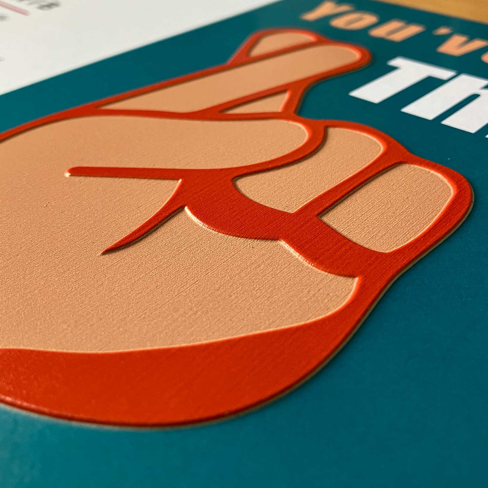 A close-up of a hand in peach and orange on a teal printed surface, with partial white text visible at the top right.