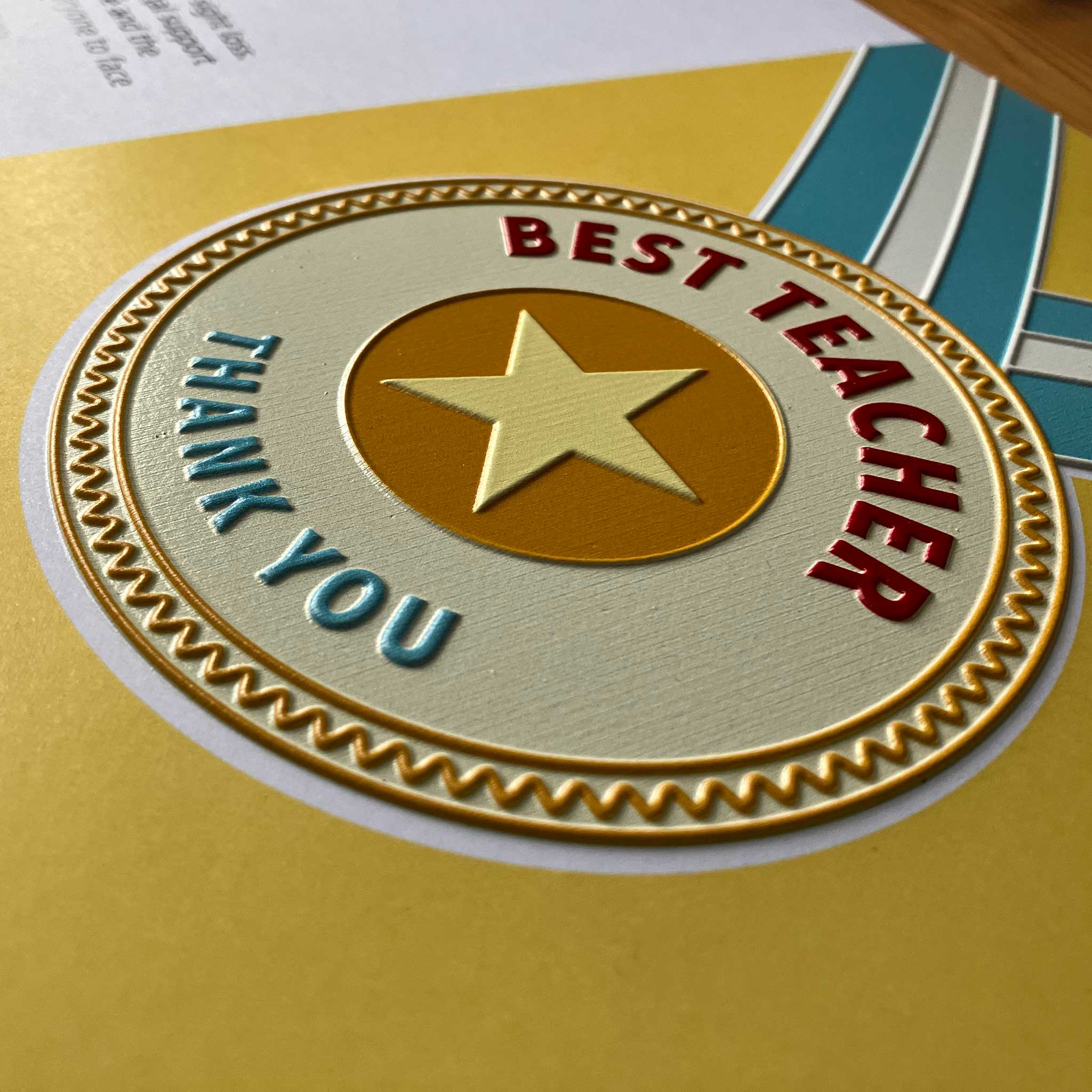 A close-up of the card or with a gold embossed star, red 'BEST TEACHER' text, and blue 'THANK YOU' text on a yellow background.
