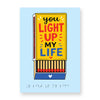Pale blue greeting card with a golden yellow matchbox carrying the message 'you light up my life' in large black, red and blue text and the drawer open to show matchsticks inside. Message repeats in braille at the bottom of the card