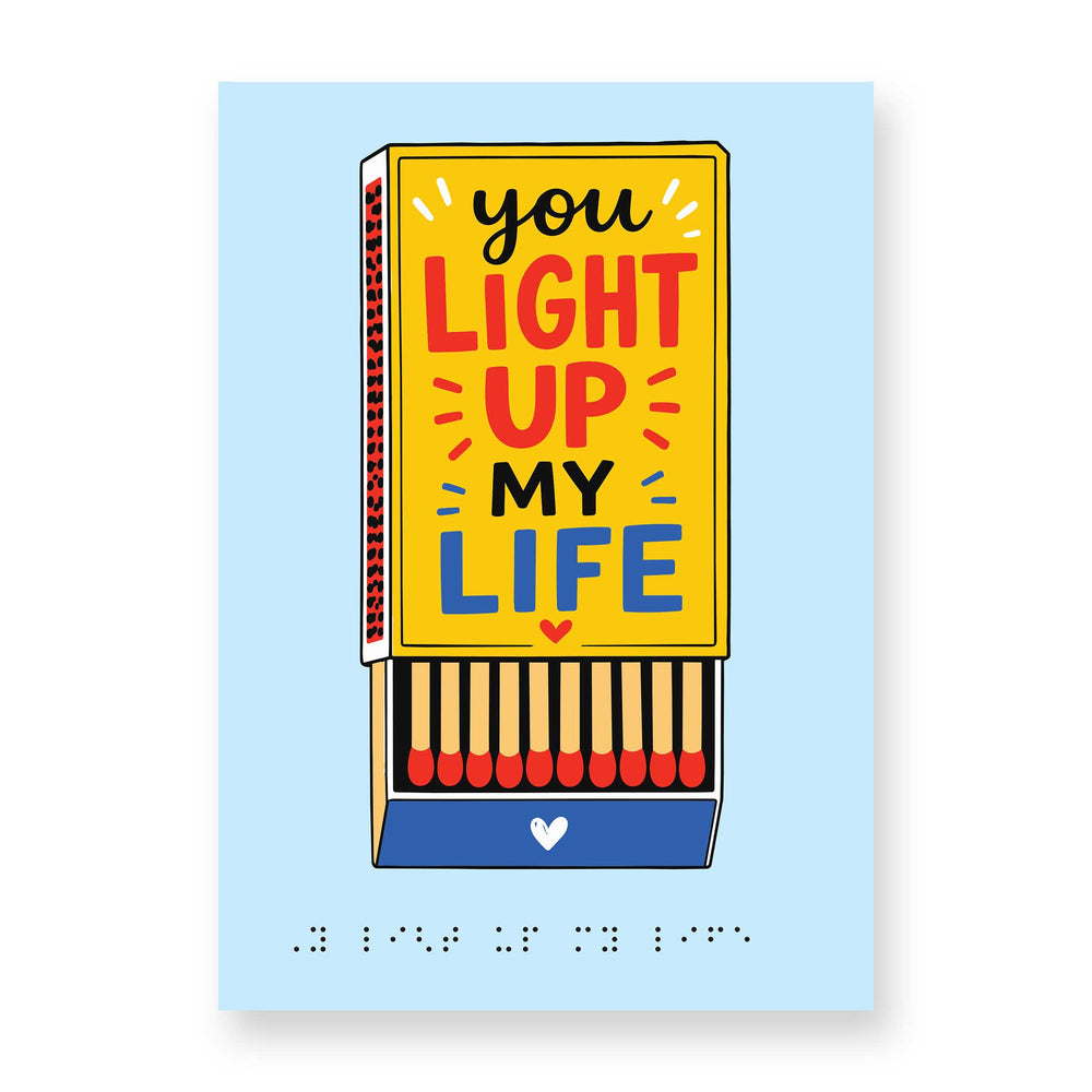 Pale blue greeting card with a golden yellow matchbox carrying the message 'you light up my life' in large black, red and blue text and the drawer open to show matchsticks inside. Message repeats in braille at the bottom of the card