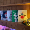 Set of Christmas-themed cards featuring a tree, reindeer, snowman, robin, and snowflake on a wooden dresser shelf lit by Christmas tree lights in a glass jar