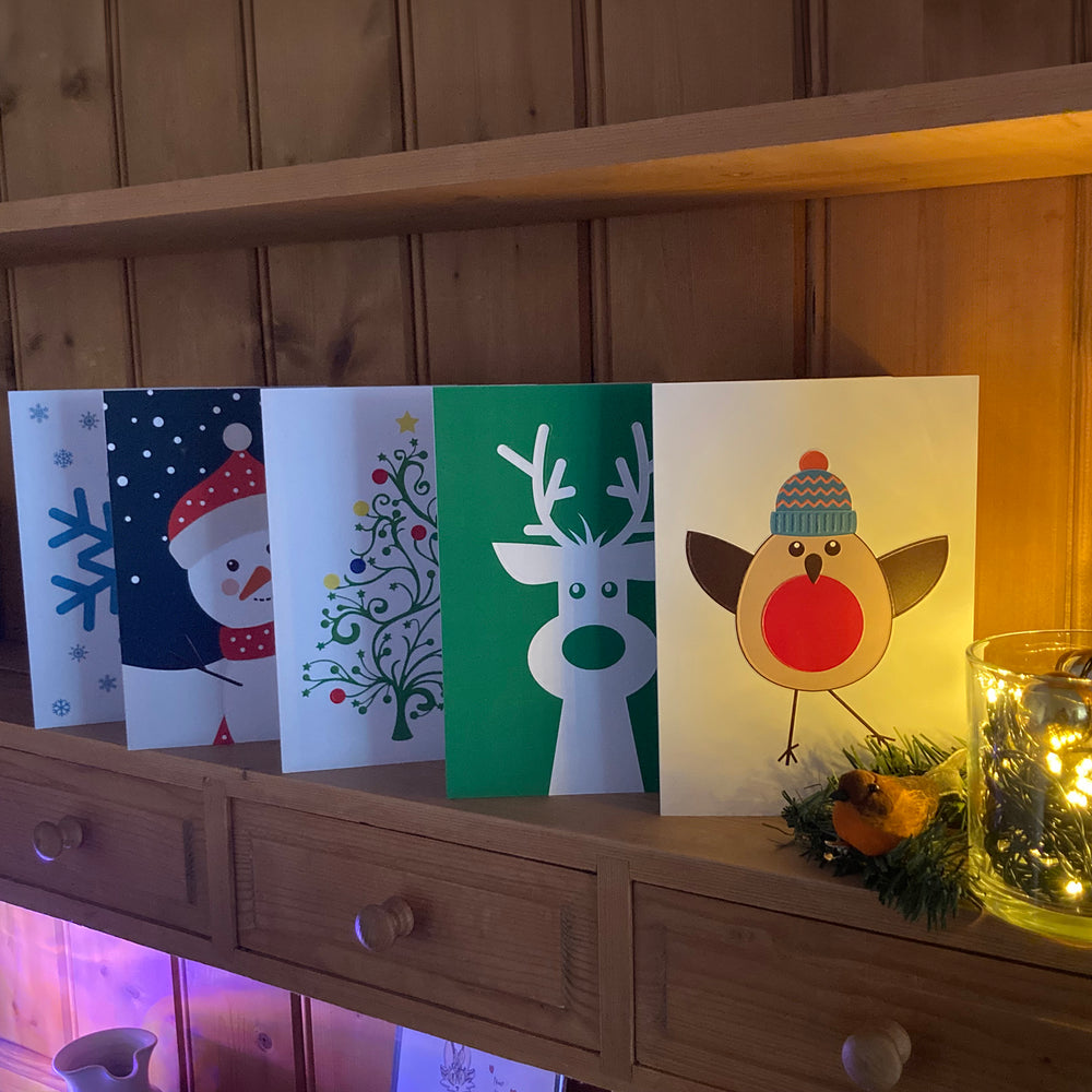 Set of Christmas-themed cards featuring a tree, reindeer, snowman, robin, and snowflake on a wooden dresser shelf lit by Christmas tree lights in a glass jar