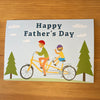 Father’s day card on a wooden desk showing a tactile image of a man and a child enjoying a tandem bicycle ride.