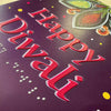 A close-up of a purple greeting card featuring raised red text that reads 'Happy Diwali' with braille underneath and colorful decorative motifs in the top right corner.