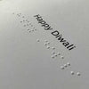 A close-up of a light gray card or paper with embossed white Braille text and the printed words 'Happy Diwali' in black at the top right.
