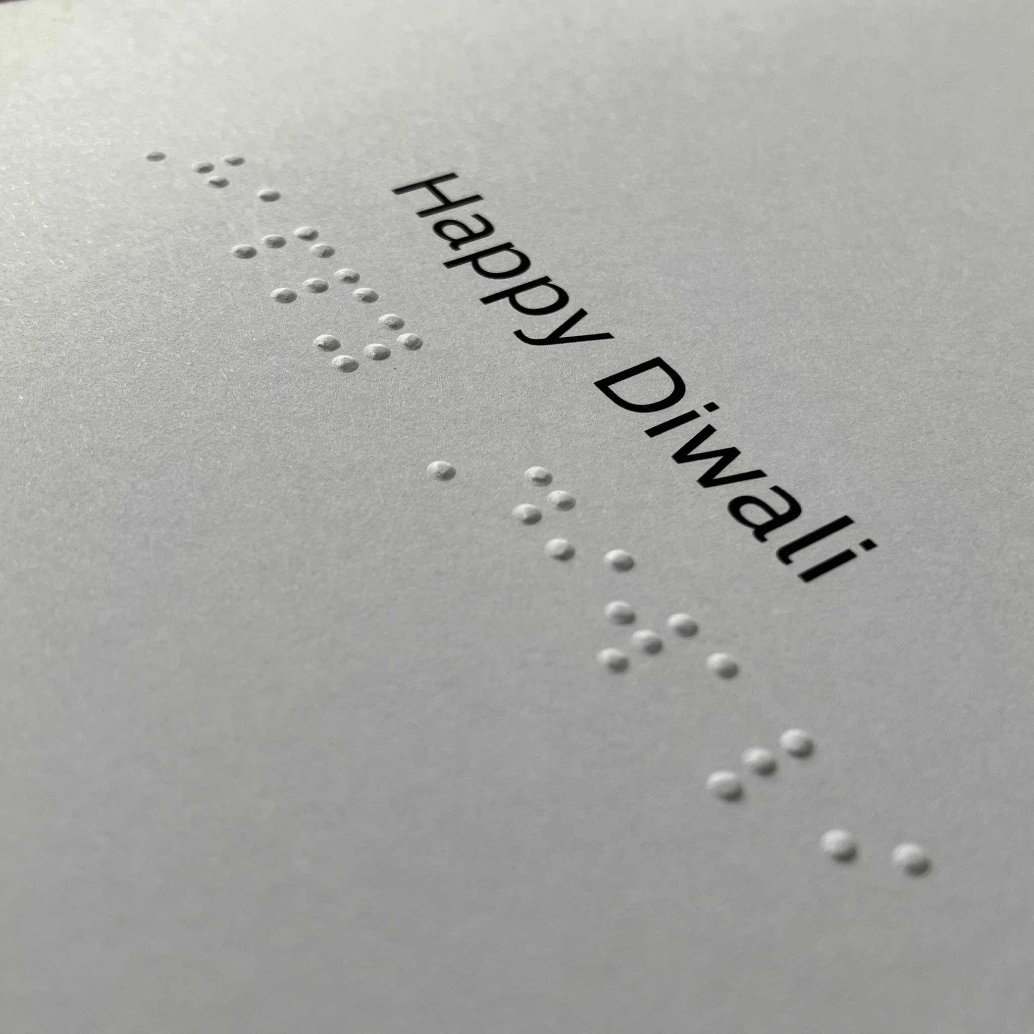 A close-up of a light gray card or paper with embossed white Braille text and the printed words 'Happy Diwali' in black at the top right.