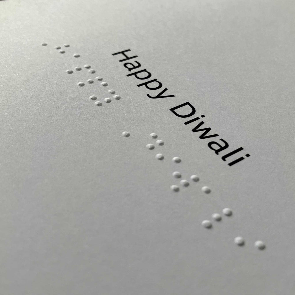 A close-up of a light gray card or paper with embossed white Braille text and the printed words 'Happy Diwali' in black at the top right.