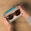 Person holding a floral-patterned glasses case with glasses inside on a wooden surface.