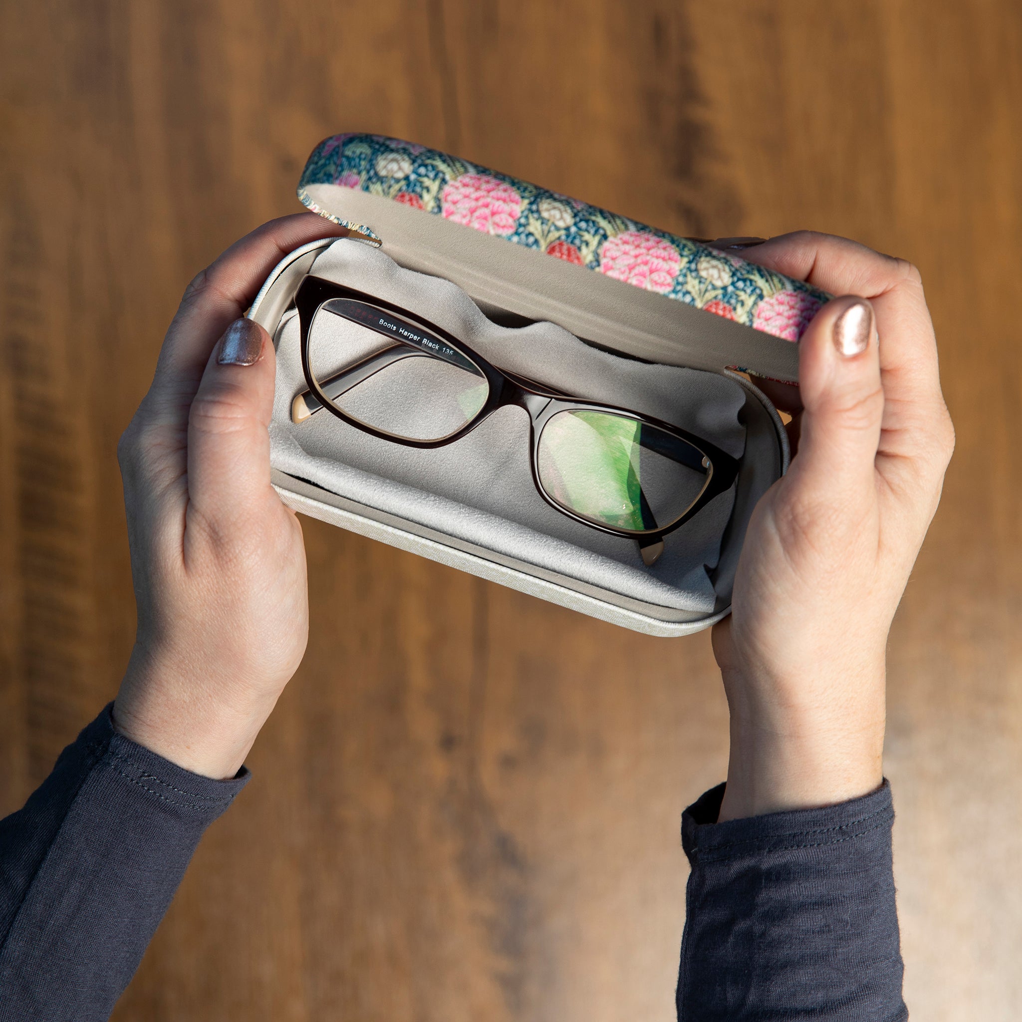 Floral-patterned glasses case held by hands against a wooden background.