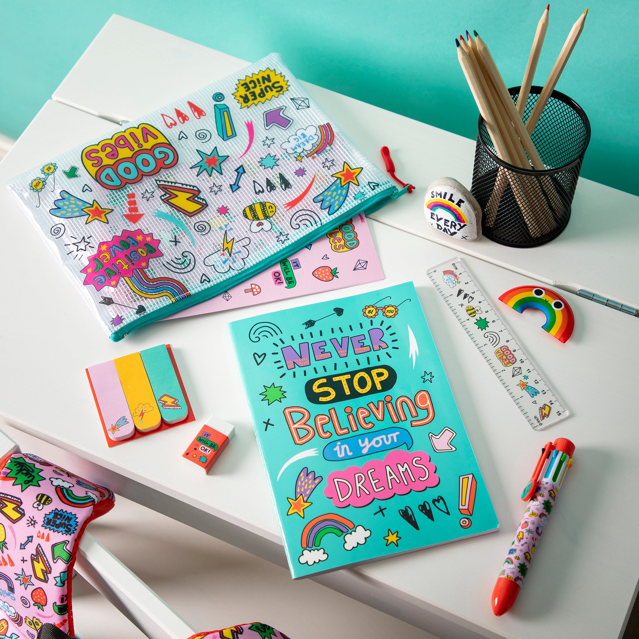 Colorful stationery items including notebooks, pens, and erasers on a white surface with a teal background.